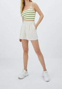 Femme portant un crop top rayé vert et blanc, un short beige taille haute, et des baskets blanches épaisses, debout avec une main dans la poche.