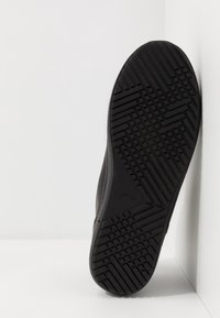 Czarna podeszwa trampka z teksturowanym wzorem bieżnika, gładkimi bokami i lekkim zakrzywieniem. Wzmocnione krawędzie zapewniają trwałość. Minimalistyczny design.