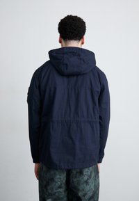 Navy blue lightweight jacket with a hood, featuring a front seam detail and a relaxed fit. Paired with patterned green pants.