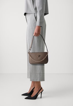 Femme en tailleur pantalon à carreaux tenant un sac taupe avec chaîne, portant des escarpins noirs à talons hauts pointus avec bride arrière, debout sur un sol blanc uni.