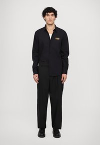 Black long-sleeve button-up shirt with a unique asymmetric design, featuring a gold logo patch on the chest. Tailored fit, paired with black trousers.