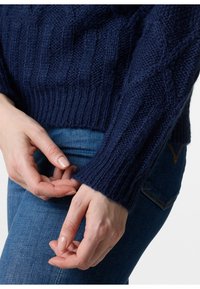 Marineblauer Strickpullover mit strukturiertem Zopfmuster, gerippten Bündchen und Saum. Kombiniert mit blauen Jeans, die den Farbkontrast des Pullovers hervorheben.