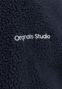 Mörk marinblå fleece tyg med vit broderad text som säger "Originals Studio" centrerad på den texturerade ytan.