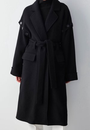 Musta villainen vyötärövyöllinen takki, jossa leveät kaulukset, käännetyt hihat, napit olkapäillä ja läppätaskut, pidetty mustan topin päällä.