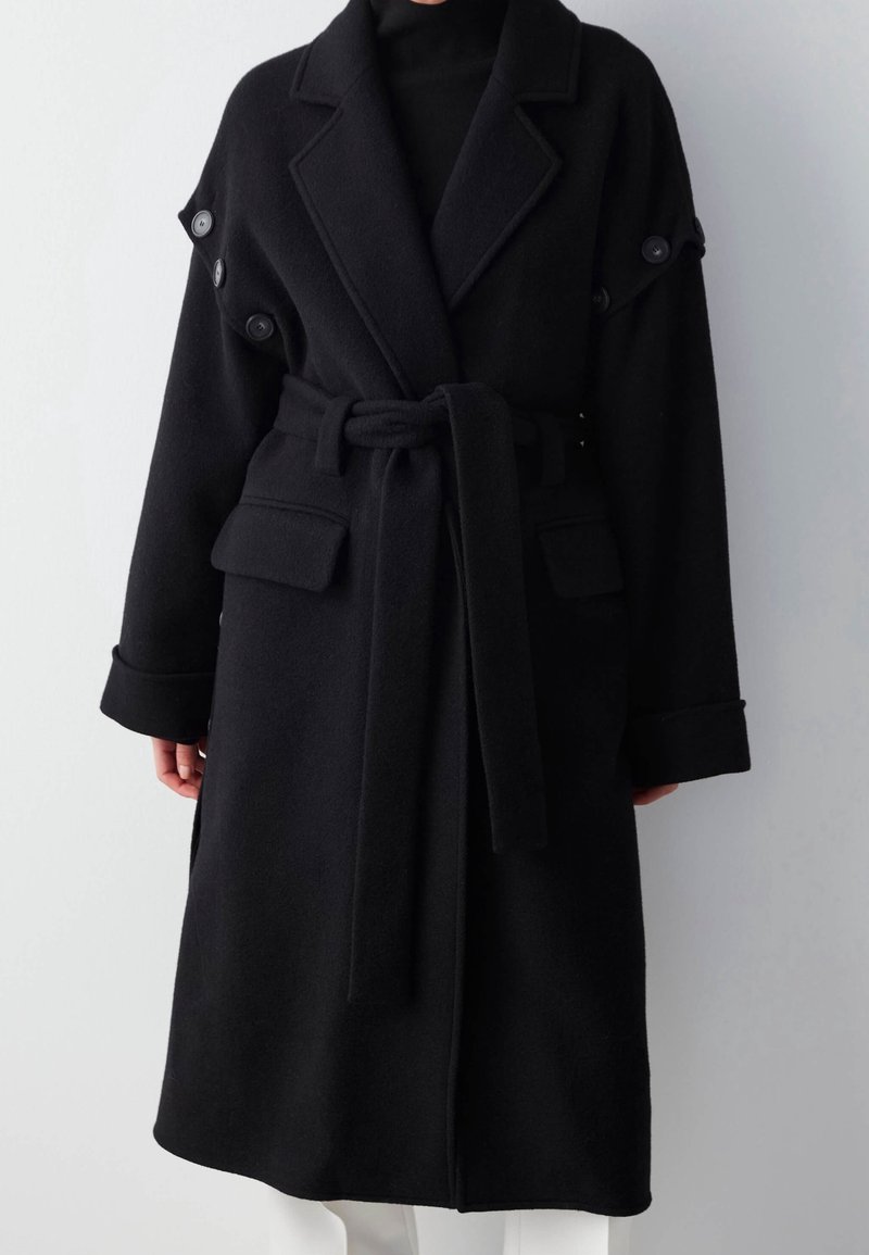 Manteau en laine noir ceinturé avec larges revers, manches retroussées, détails de boutons aux épaules et poches à rabat porté sur un top noir.
