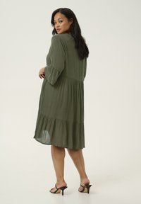 Vestido verde oliva de corte holgado con mangas tres cuartos, dobladillo con volantes y tejido texturizado, combinado con tacones negros de tiras.