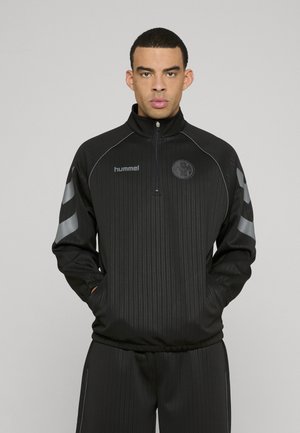 Veste noire zippée avec des rayures verticales, comportant des accents gris et un logo sur la poitrine. Le matériau semble léger et sportif.