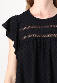 Blouse noire à manches courtes froncées, ornée de points texturés et d'un panneau en dentelle sur la poitrine, en tissu doux, coupe décontractée.