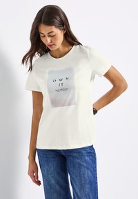 Femme portant un t-shirt blanc avec le texte "OWN IT" et un jean bleu, regardant vers le bas avec une main derrière le dos, sur fond blanc uni.