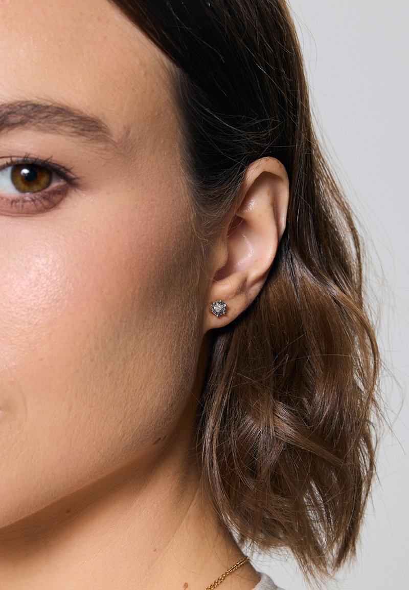 Srebrna kolczyk z przezroczystym kamieniem w kształcie okrągłym, na tle gładkiej, jasnej karnacji i włosów brązowych do ramion.