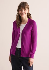 Cardigan violet vif en tissu doux, avec un col en V, une fermeture à boutons sur toute la longueur et des détails côtelés aux poignets et à l'ourlet, porté sur une chemise blanche.