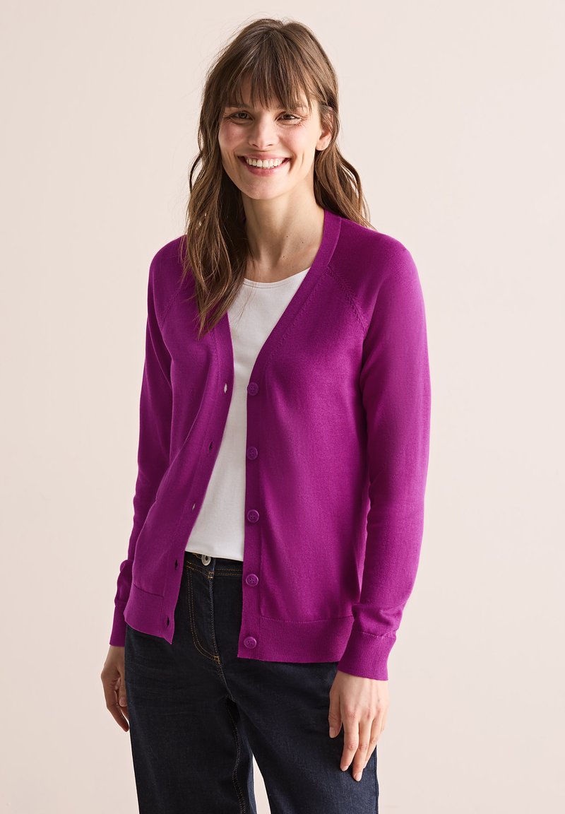 Cardigan violet vif en tissu doux, avec un col en V, une fermeture à boutons sur toute la longueur et des détails côtelés aux poignets et à l'ourlet, porté sur une chemise blanche.