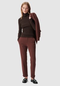 Maglione a collo alto marrone a coste abbinato a pantaloni tapered bordeaux e mocassini neri, accessoriato con uno strato esterno abbinato appoggiato su un braccio.