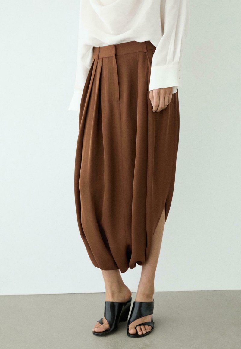 Brązowe szerokie spodnie plisowane z marszczonymi mankietami, w połączeniu z czarnymi sandałami na obcasie z odkrytymi palcami oraz białą koszulą z długim rękawem.