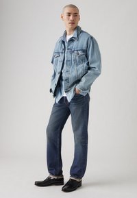 Chaqueta de mezclilla azul claro con un ajuste relajado, cierre de botones, bolsillos en el pecho, combinada con jeans azul oscuro y zapatos de cuero negros.