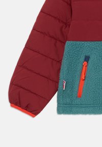 Red and teal fleece jacket with a puffy design, textured body, and an orange zip pull. Features an attached logo tag and elastic cuffs.