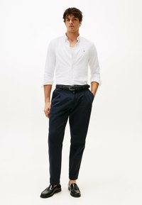 Camicia bianca con bottoni, chinos blu navy, cintura intrecciata nera e mocassini neri. La camicia ha un colletto classico, i chinos presentano tasche laterali.