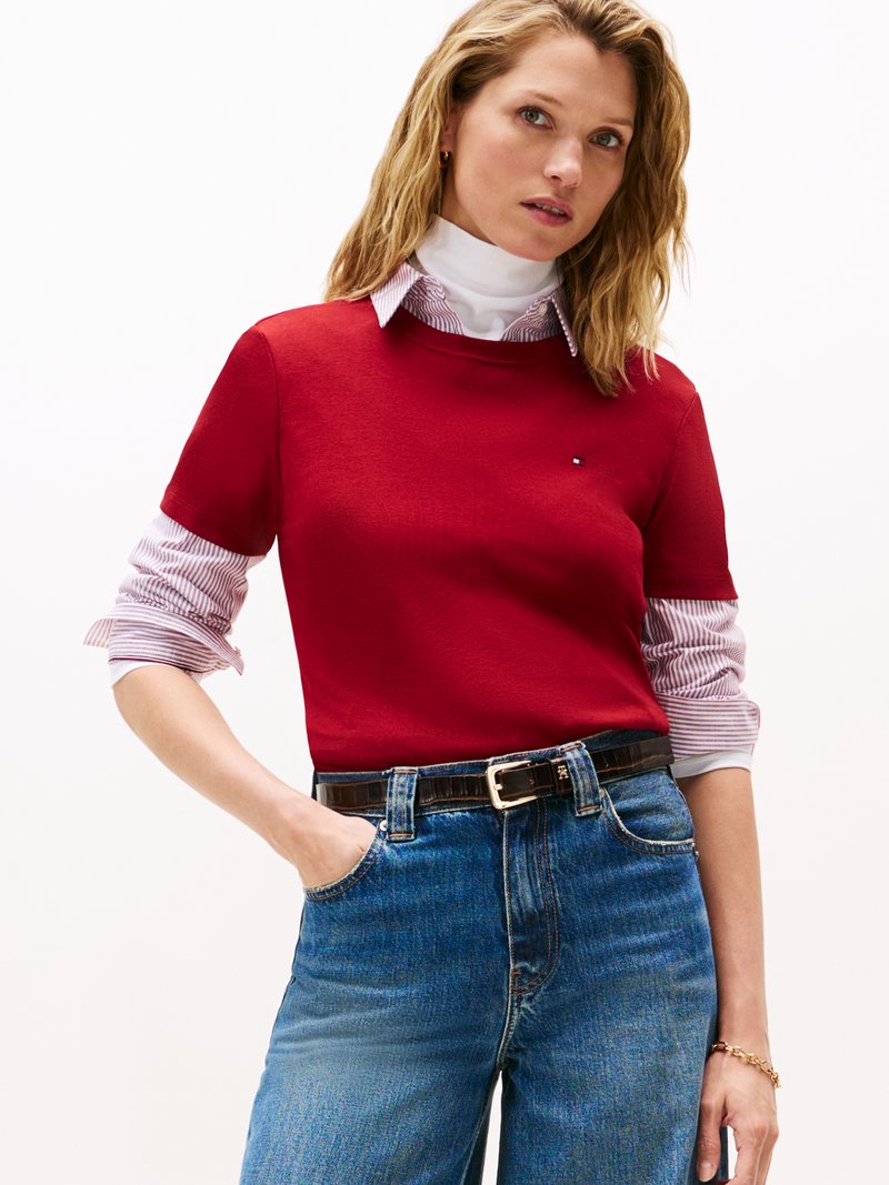 Camiseta de algodón roja superpuesta sobre una camisa de cuello blanca con rayas rojas, combinada con jeans azules y un cinturón oscuro, con un pequeño detalle de logo.