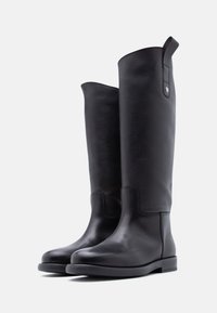 Ψηλές μαύρες δερμάτινες μπότες με λεία υφή, στρογγυλεμένα δάχτυλα, ελάχιστες ραφές και μικρό βοηθητικό λουράκι στο πάνω μέρος για εύκολη εφαρμογή.