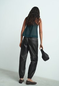 Crne hlače od umjetne kože s elastičnim manšetama, uparene s tirkiznim prozirnim topom na bretele. Osoba drži malu crnu pletenu torbu.