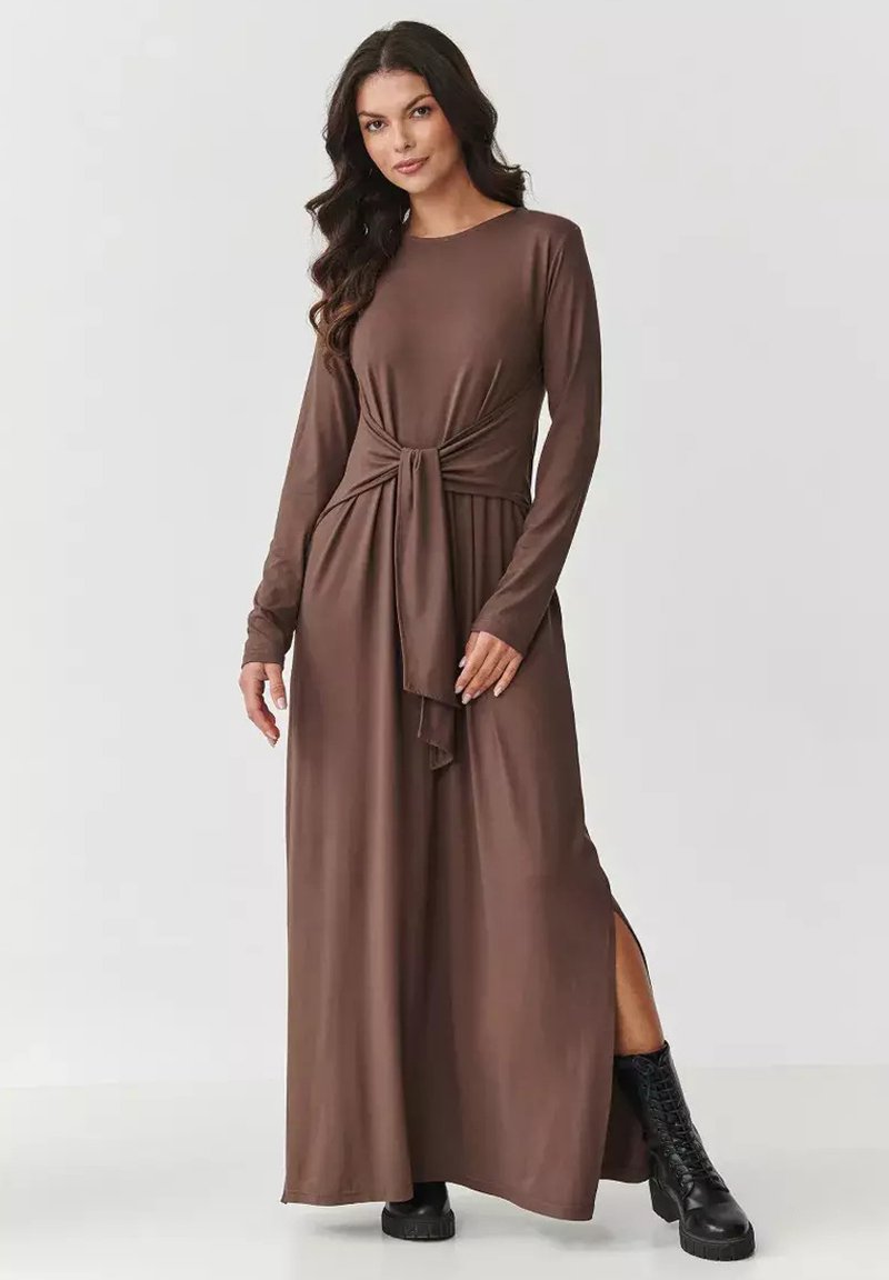 Brązowa, długa sukienka maxi z długim rękawem, z węzłem w talii i bocznym rozcięciem; wykonana z miękkiego materiału, zestawiona z czarnymi, sznurowanymi butami.