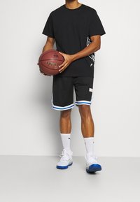 Černé tričko s potiskem, černé síťované šortky s modrými a bílými akcenty a bílé tenisky s modrou podrážkou, držící basketbal.