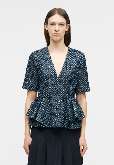 Femme portant une veste péplum texturée bleu marine à manches courtes, avec cinq boutons sur le devant et un col en V, associée à une jupe plissée foncée.