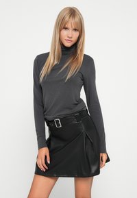 Sivi dugi rukav turtleneck top u kombinaciji s crnom suetnom mini suknjom s plitvicama koja ima detalj remena i kopče. Glatka tekstura, kroj koji se prilagođava tijelu.