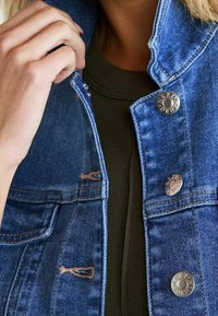 Farkkutakki haalistuneella sinisellä värillä, näkyvillä saumoilla ja hopeisilla painonapeilla. Siinä on korkea kaulus ja kulunut pinta.