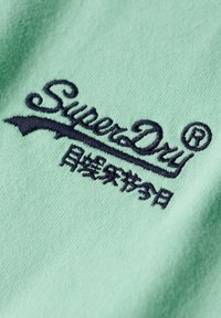 Mintgrøn stof med en blød texture, der har et mørkeblåt broderet logo "SuperDry" og yderligere tekst nedenfor.