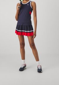 Marinblå linne med röda detaljer, kombinerad med en veckad kjol med röda och vita ränder. Modellen bär marinblå skor och vita ankelsockor.