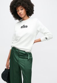 Νεαρή γυναίκα με σγουρά μαλλιά, ντυμένη με λευκό φούτερ Nike και πράσινο παντελόνι, κρατώντας ένα μαύρο καπέλο στο αριστερό της χέρι.