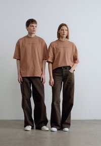 Καφέ φαρδιά μπλούζα με μικρό γραφικό, σε συνδυασμό με καφέ παντελόνια με φαρδιά πόδια. Λευκά αθλητικά παπούτσια ολοκληρώνουν την μίνιμαλ, καθημερινή εμφάνιση.