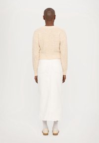 Personne à la tête rasée vue de dos, portant un pull beige texturé, une jupe midi blanche, des chaussettes blanches et des chaussures beiges.