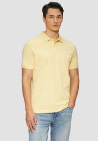 Polo en coton jaune clair avec un col et une patte de boutonnage à deux boutons. Porté avec un jean en denim bleu, avec un logo subtil sur la poitrine.