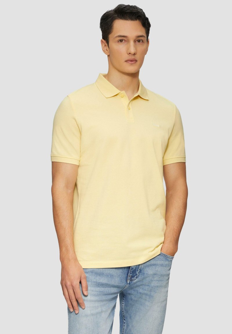 Polo en coton jaune clair avec un col et une patte de boutonnage à deux boutons. Porté avec un jean en denim bleu, avec un logo subtil sur la poitrine.