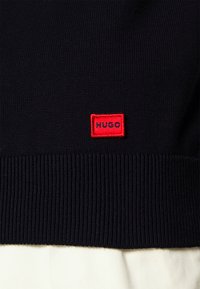 Navy knitted sweater with ribbed cuffs; features a prominent red rectangular label with "HUGO" in black lettering at the hem.
