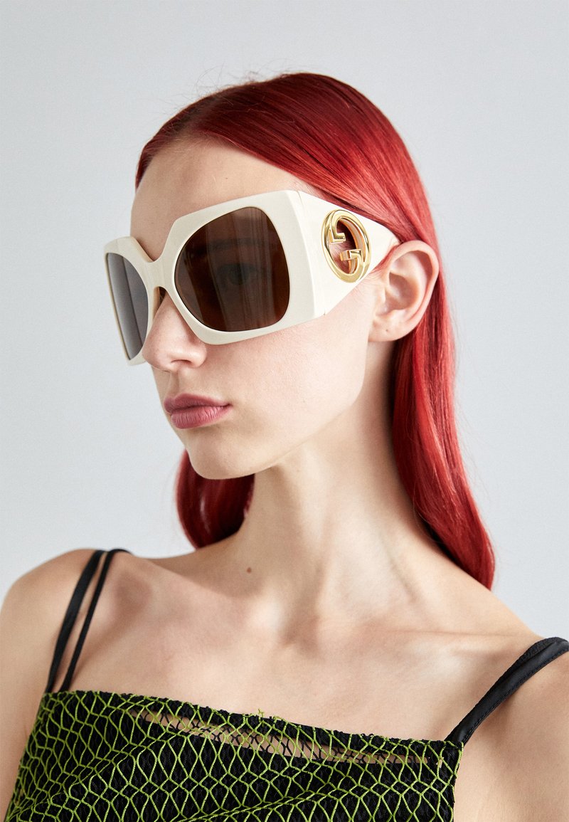 Gafas de sol blancas de gran tamaño de Gucci con lentes marrones, que presentan un distintivo logotipo dorado en el lateral. Material suave y diseño geométrico audaz.