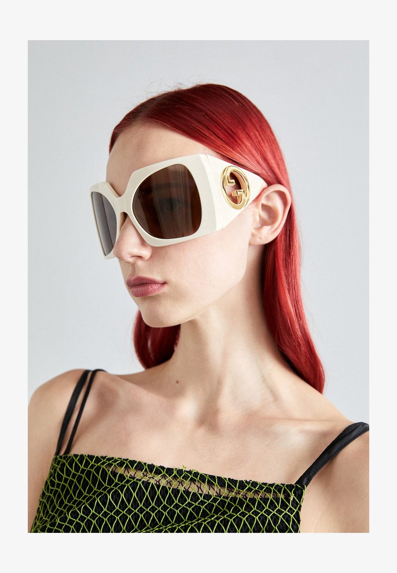 Přesměrované bílé sluneční brýle Gucci s hnědými skly, které mají výrazné zlaté logo na boku. Hladký materiál a odvážný geometrický design.