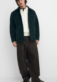 Giacca camicia in pile verde con due tasche sul petto, indossata sopra una camicia crema con colletto e pantaloni marroni a gamba larga con motivo. Scarpe scure completano il look.