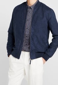 Námořnická modrá bomber bunda s hladkým semišovým povrchem, žebrovanými manžetami a límcem, zipovým zapínáním, nošená přes vzorovanou košili a bílé kalhoty.