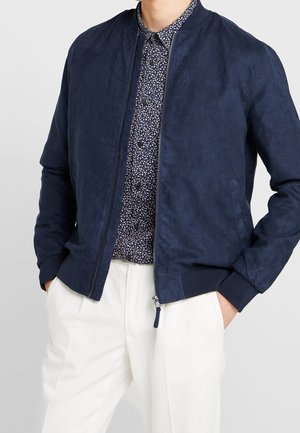 Mörkblå bomberjacka med en slät mockaliknande yta, ribbade manschetter och krage, dragkedja framtill, bärs över en mönstrad skjorta och vita byxor.