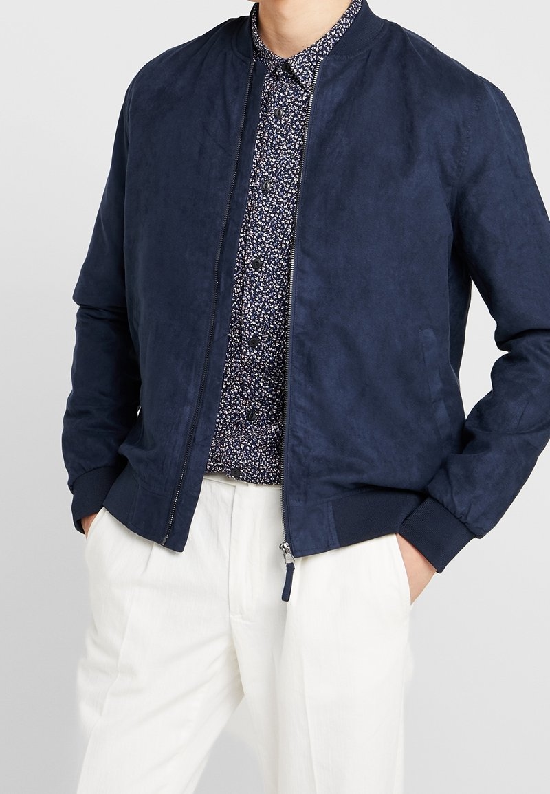 Námořnická modrá bomber bunda s hladkým semišovým povrchem, žebrovanými manžetami a límcem, zipovým zapínáním, nošená přes vzorovanou košili a bílé kalhoty.