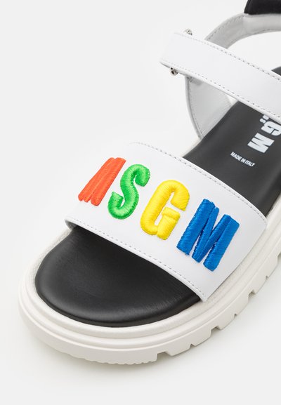 Sandale en cuir blanc avec une épaisse semelle noire. Présente des lettres brodées colorées "MSG" en rouge, vert, jaune et bleu sur la sangle.