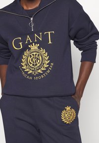 Sudadera navy con media cremallera, que presenta un logotipo de GANT en oro y detalle de corona. Combinada con pantalones a juego que muestran un emblema similar.