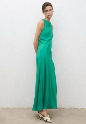 Woman wearing a sleeveless, floor-length green dress and beige heeled sandals, standing against a plain white wall with hands behind her back.