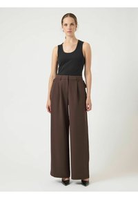 Donna in piedi, che indossa un top senza maniche nero, pantaloni marroni a vita alta e gamba larga, e scarpe nere a punta, su uno sfondo semplice.