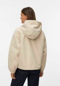 Sieviete ar gariem brūniem matiem, ģērbusies bēša krāsas kapucētas jakā un tumšās džinsās, redzama no muguras, baltā vienkāršā fona priekšā.