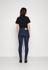 T-shirt preto de manga curta combinado com jeans de ganga azul escura de cintura alta. Os jeans possuem dois bolsos traseiros e um rótulo em simulação de pele castanha. Botins pretos.