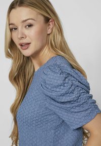 Modrá textúrovaná blúzka s vypukanými krátkymi rukávmi a okrúhlym výstrihom. Materiál má bublinkový vzor, ktorý zvyšuje vizuálny záujem.